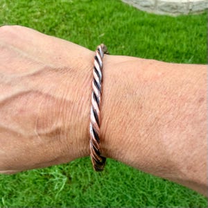 May include: A twisted copper bracelet with a braided design. The bracelet is a mix of copper and silver tones, creating a striking contrast. The jewelry is worn on a wrist, set against a backdrop of green grass.