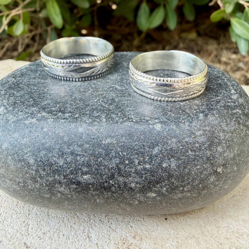 May include: Two silver rings with a textured band design. Each ring features a central row of repeating diamond-shaped patterns bordered by thin, beaded edges. The metal has a polished finish with darkened recessed areas to highlight the details.