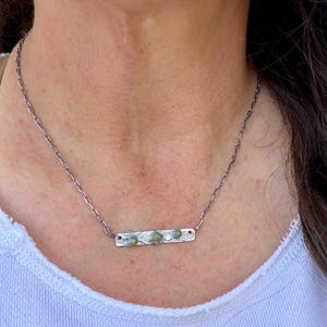 May include: A silver-toned metal necklace featuring a rectangular bar pendant with an etched leaf pattern. The bar is set with three small, round, green-toned stones and one central square-cut green stone, suspended from a delicate chain.