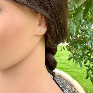 May include: A close-up of a person's ear with a small, round stud earring. The earring features a light blue gemstone set in a silver or white metal setting. The background is blurred, with green foliage visible.