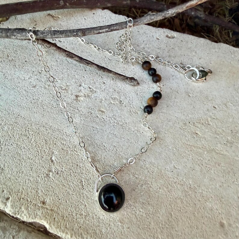 May include: A silver chain necklace featuring a single round black stone pendant in a silver setting. The chain includes a small section of alternating black and brown spherical beads near the clasp, which is a standard silver lobster claw style.