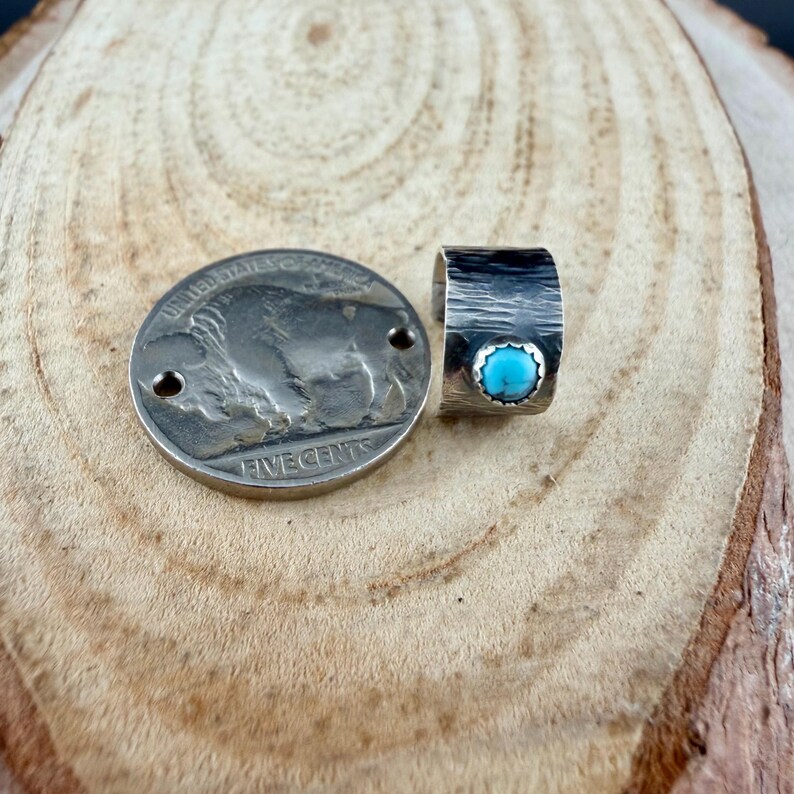 May include: A silver-toned ring with a textured band and a small, round, turquoise stone. Next to the ring is a United States nickel coin. The ring and coin are displayed on a wooden surface.