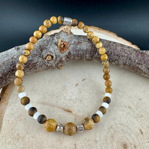 May include: A beaded bracelet featuring a mix of earth-toned jasper, tiger's eye, and white beads. The bracelet is strung on a flexible cord and secured with a silver-toned clasp. The beads vary in size and texture, creating a natural, organic look.