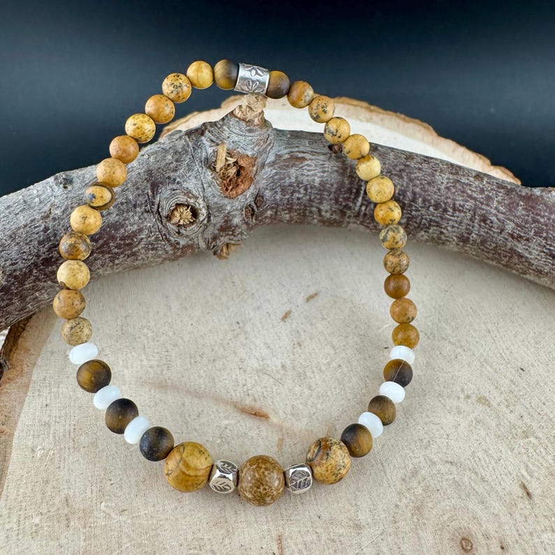 May include: A beaded bracelet featuring a mix of earth-toned jasper, tiger's eye, and white beads. The bracelet is strung on a flexible cord and secured with a silver-toned clasp. The beads vary in size and texture, creating a natural, organic look.