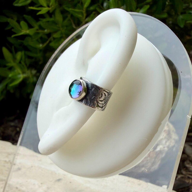May include: A silver ring with a round, iridescent gemstone. The band features an etched design. The ring is displayed on a white hand-shaped form, with a blurred green background.