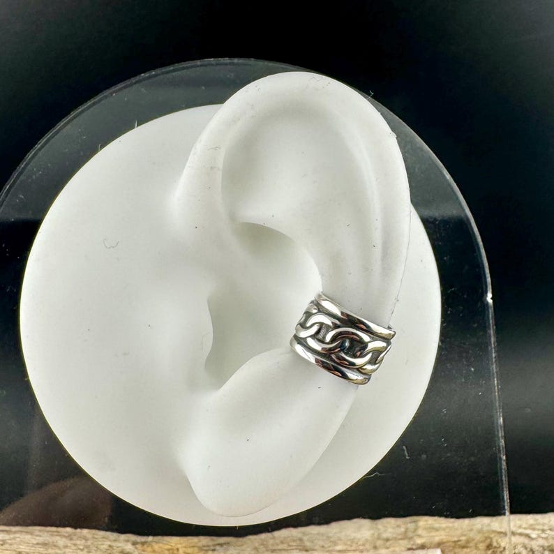 May include: A silver ear cuff with a chain link design. The cuff is shown on a white ear model. The ear cuff is a non-pierced earring that wraps around the ear. The jewelry is made of a shiny, metallic material.