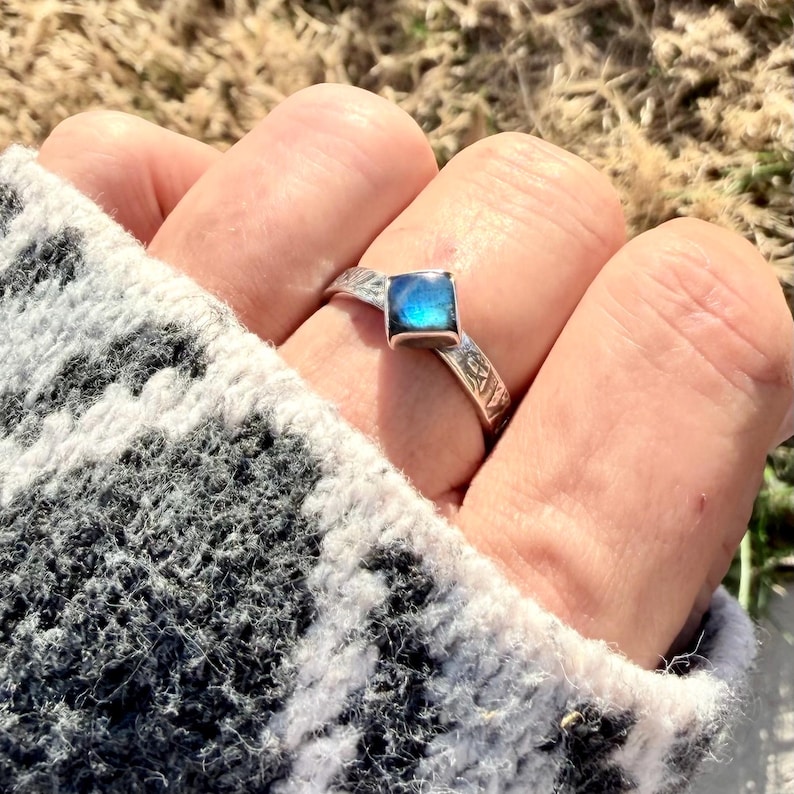 May include: A silver ring with a square, iridescent blue gemstone. The ring band features an etched design. The ring is worn on a finger, with a gray and white patterned sleeve in the background.