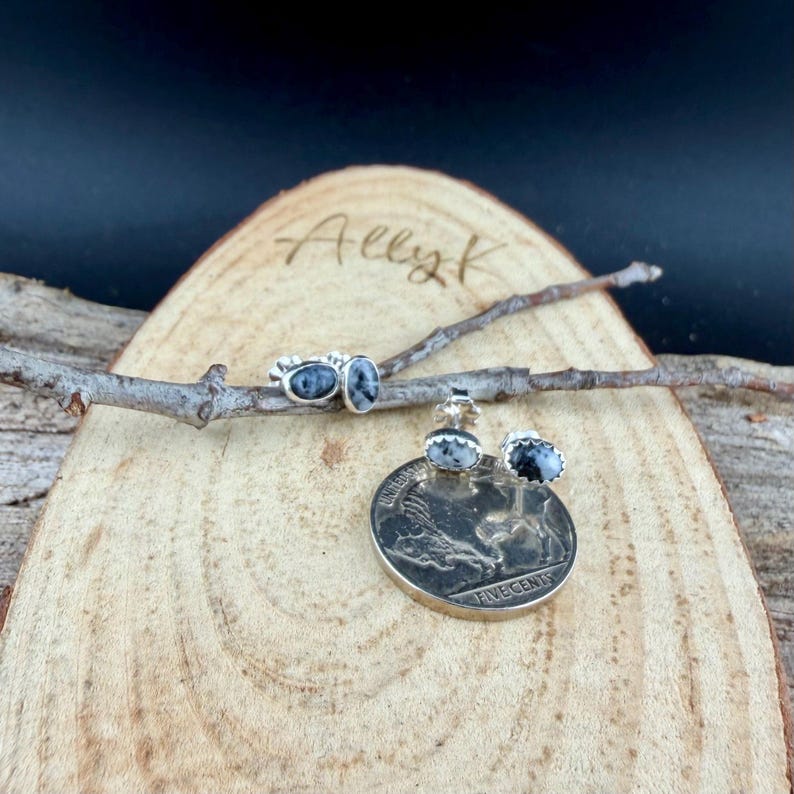 May include: A jewelry set featuring a ring, pendant, and earrings. The ring and earrings have oval-shaped stones with a black and white pattern. The pendant is a silver coin with a buffalo design. The jewelry is displayed on a wooden surface.