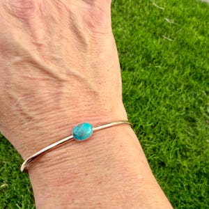 May include: A gold-toned cuff bracelet featuring a vibrant, oval turquoise stone. The bracelet is simple in design, with a slender band that curves around the wrist. The turquoise stone is the focal point, adding a pop of color against the gold.