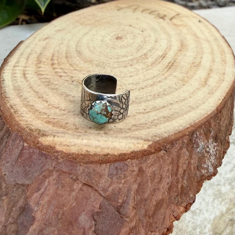 May include: A silver-toned ring with a turquoise stone. The ring has a wide band with etched patterns. The turquoise stone is oval-shaped and set in a bezel. The ring is displayed on a wooden surface.