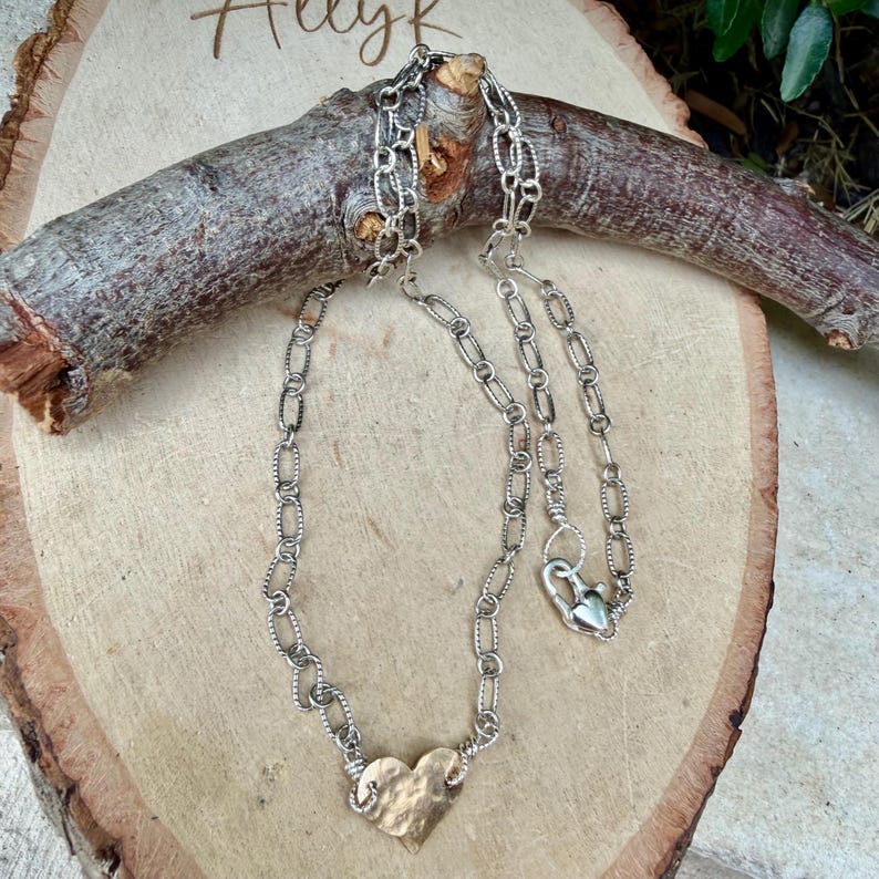 May include: A silver-toned chain necklace featuring a textured, heart-shaped pendant in a metallic gold finish. The necklace is constructed with elongated oval links and includes a lobster claw clasp for closure, showcasing a mixed-metal design.