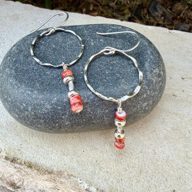 May include: A pair of silver hoop earrings featuring a hammered texture. Each earring has a dangling charm composed of two small, round, coral-colored beads separated by a silver spacer bead, suspended from the bottom of the circular hoop frame.