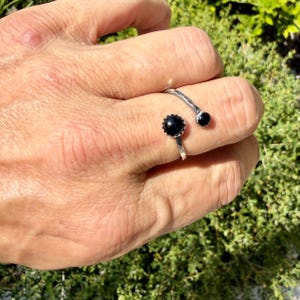 May include: An open-style silver ring featuring two black circular stones of different sizes. The larger stone is set in a scalloped bezel, while the smaller stone is set in a simple round bezel, connected by a textured, slightly hammered silver band.
