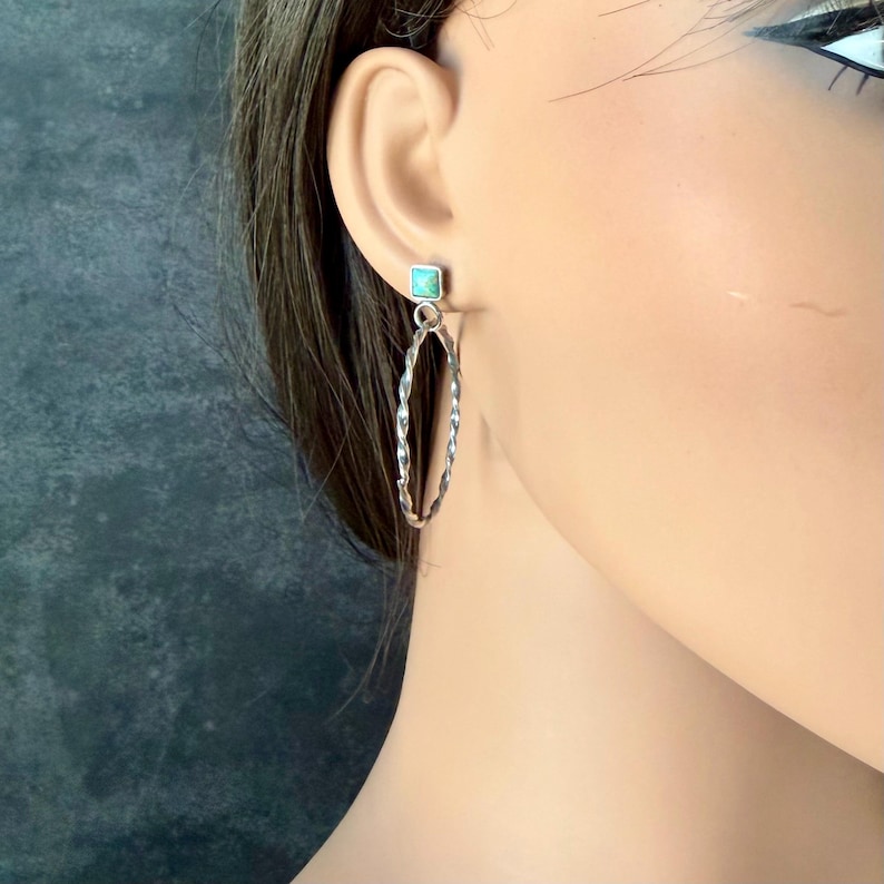 May include: Silver-toned hoop earrings with a twisted design. Each earring features a small, square turquoise stone at the top. The earrings are shown on a mannequin, highlighting their shape and detail.
