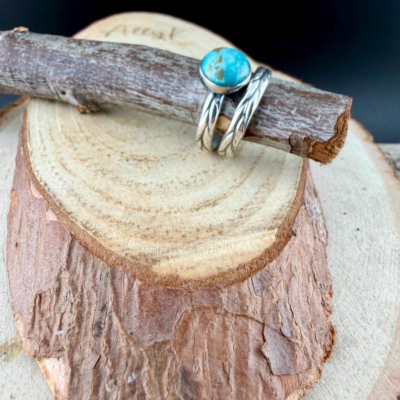 May include: A silver ring with a turquoise stone, resting on a wooden branch. The ring has a twisted band design. The turquoise stone is round and light blue. The ring is displayed on a wooden slice with a natural bark edge.