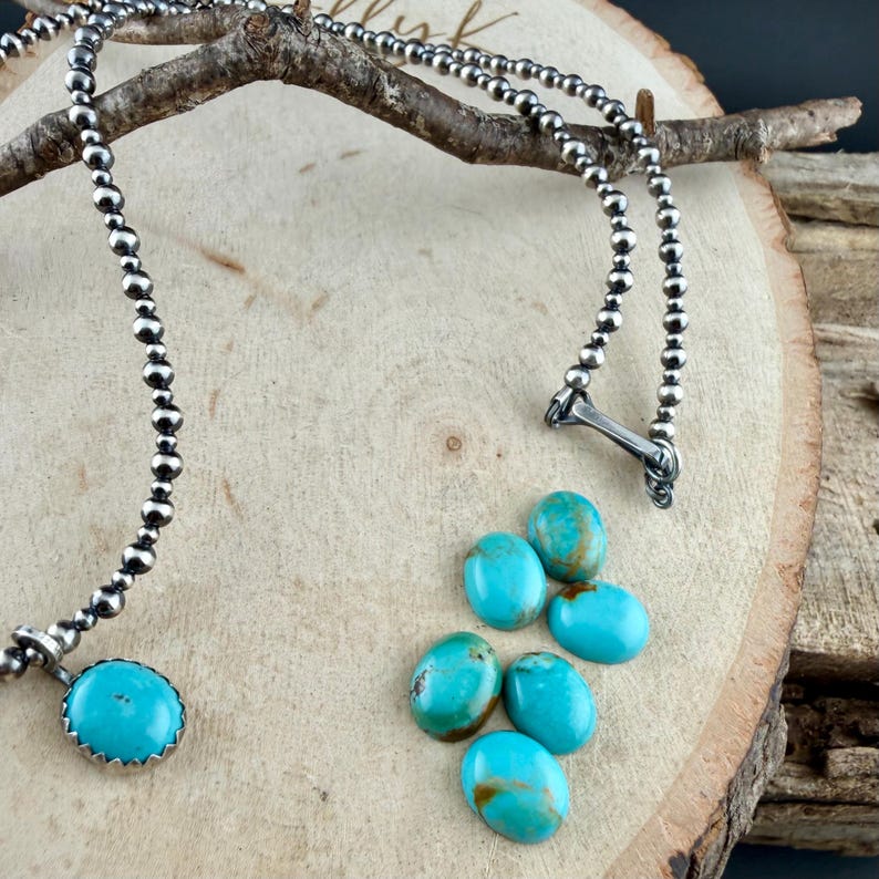 May include: A silver beaded necklace with a turquoise pendant and matching turquoise stones. The necklace is displayed on a wooden surface with a branch. The pendant is round with a silver bezel setting. The turquoise stones are oval-shaped.