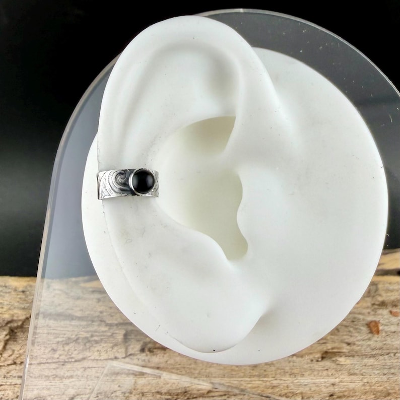 May include: A silver ear cuff with a black gemstone. The cuff features an engraved design and is shown on a white ear model. The jewelry is displayed against a dark background.