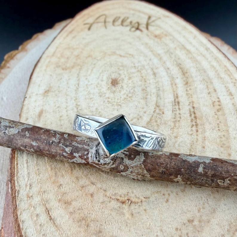 May include: A silver ring with a square, dark blue gemstone. The band has an etched design. The ring is displayed on a wooden branch against a wood slice background. The text "Ally K" is visible on the wood slice.