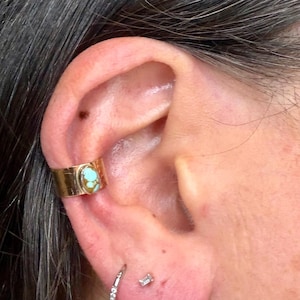 May include: Gold-colored ear cuff with a turquoise stone set in the center. The cuff is worn on the upper ear, and a small diamond earring is visible in the earlobe.