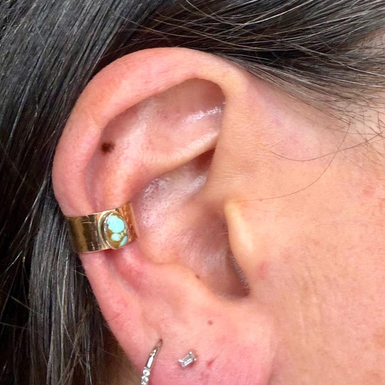 May include: Gold-colored ear cuff with a turquoise stone set in the center. The cuff is worn on the upper ear, and a small diamond earring is visible in the earlobe.
