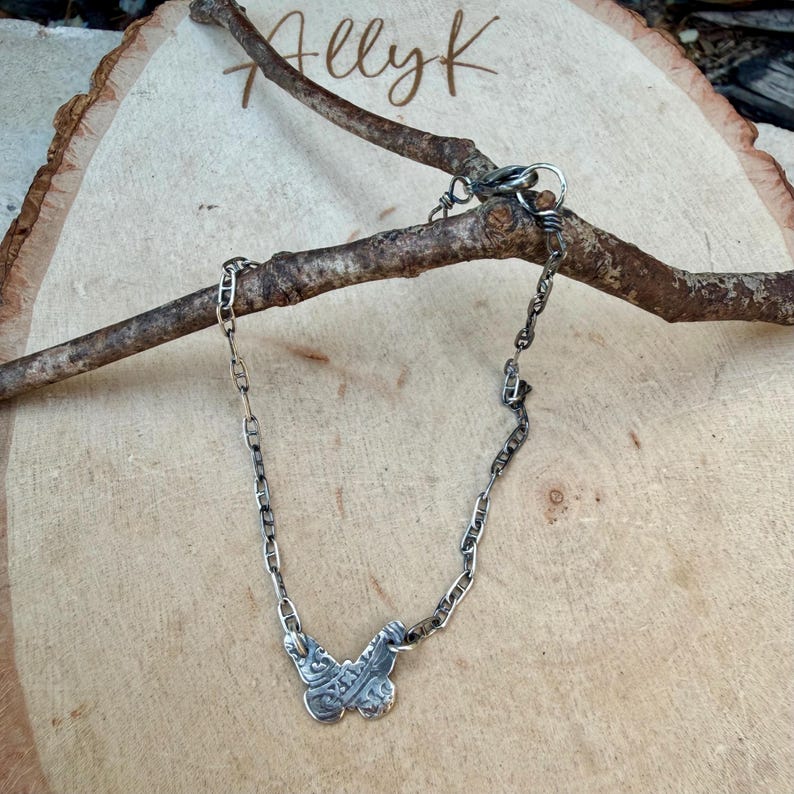 Op de afbeelding: Een zilverkleurige ketting met een vlinderhanger. De vlinderhanger heeft ingewikkelde details. De ketting wordt gepresenteerd op een houten oppervlak met de handgeschreven tekst "Ally K". De ketting is over een bruine tak gedrapeerd.