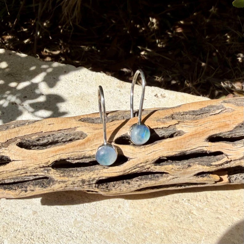 May include: A pair of silver drop earrings featuring round, translucent moonstone cabochons. Each earring has a curved silver wire hook that attaches to the circular stone setting, showcasing a subtle blue iridescent glow within the smooth surface.