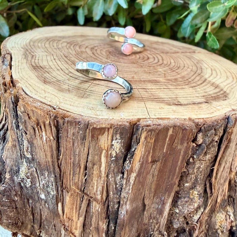 May include: Two silver rings with open bands, each featuring two round, light pink cabochon stones set at the ends. The metal bands have a smooth, polished finish, and the stones are securely held in place by small, circular silver settings.