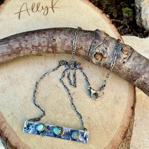 May include: A silver necklace with a rectangular pendant featuring three turquoise stones. The pendant has a textured, hammered metal design. The chain is a linked style with a lobster clasp. The name "Allyk" is written in cursive in the upper left corner.