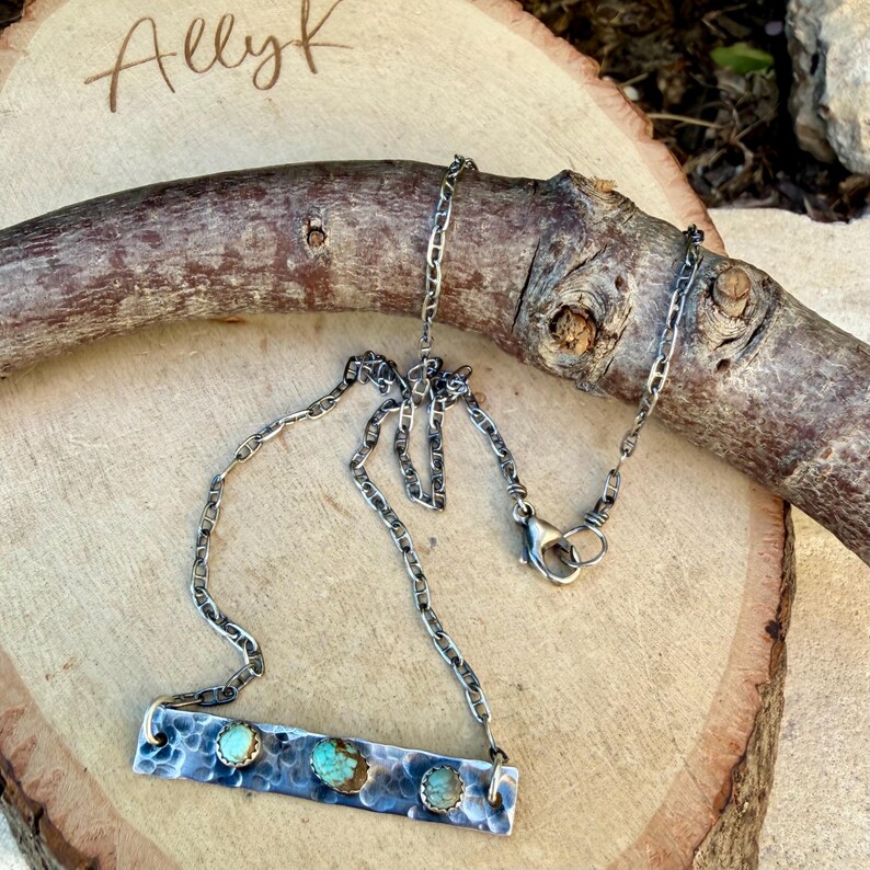 May include: A silver necklace with a rectangular pendant featuring three turquoise stones. The pendant has a textured, hammered metal design. The chain is a linked style with a lobster clasp. The name "Allyk" is written in cursive in the upper left corner.