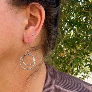 May include: A silver-toned dangle earring featuring two concentric circles. The outer circle has a twisted, rope-like texture, while the inner circle is smooth and plain. The earring hangs from a thin, simple silver wire hook.