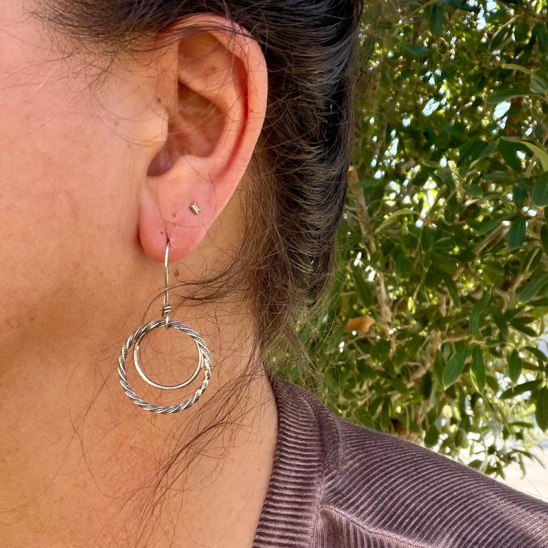 May include: A silver-toned dangle earring featuring two concentric circles. The outer circle has a twisted, rope-like texture, while the inner circle is smooth and plain. The earring hangs from a thin, simple silver wire hook.
