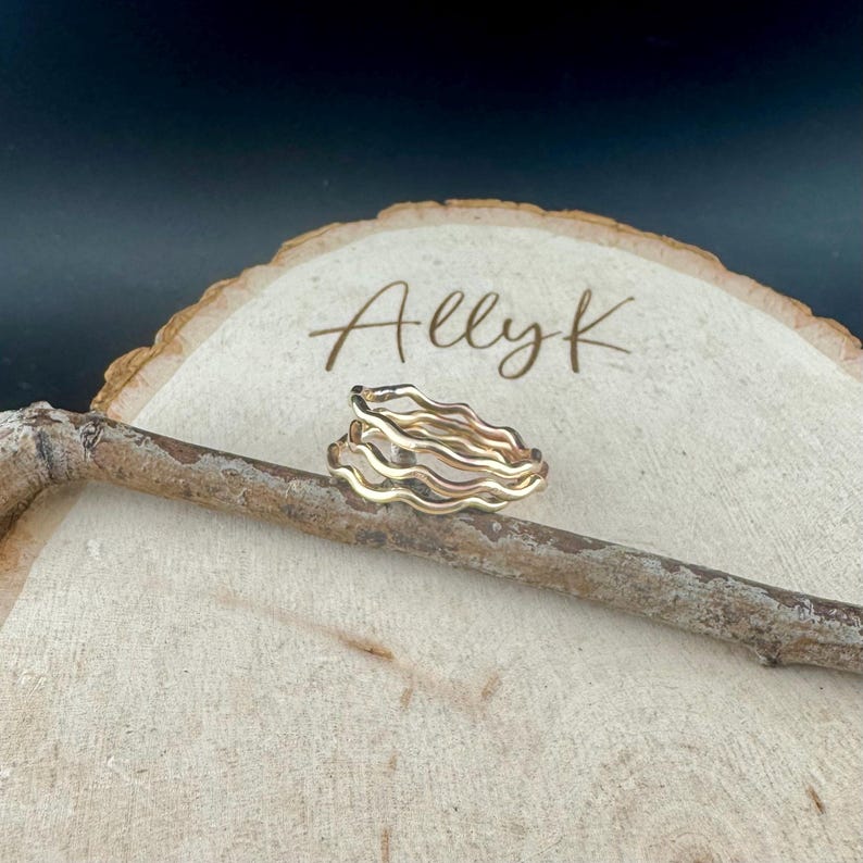 May include: A gold-toned ring with a wavy design, resting on a wooden surface. The ring has multiple layers, creating a textured appearance. The word "Allyk" is written on the wooden surface.