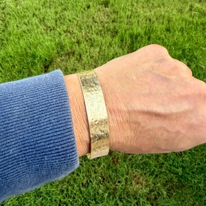 Puede incluir: Un brazalete tipo pu&ntilde;o de metal en tono dorado con una superficie texturizada y martillada. La banda ancha se lleva en la mu&ntilde;eca, mostrando un brillo met&aacute;lico constante y un patr&oacute;n irregular repetido en toda la superficie exterior de la joya.