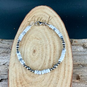 May include: A beaded bracelet featuring white and gray stone beads interspersed with silver-colored metal beads. The bracelet is strung on a flexible cord and has a silver-colored clasp. The bracelet is displayed on a wooden surface.
