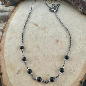 May include: A silver necklace with black beads. The necklace features a delicate chain design with evenly spaced, round black beads. The beads are interspersed with small silver accents, creating a contrast against the dark beads. The necklace is displayed on a wooden surface.