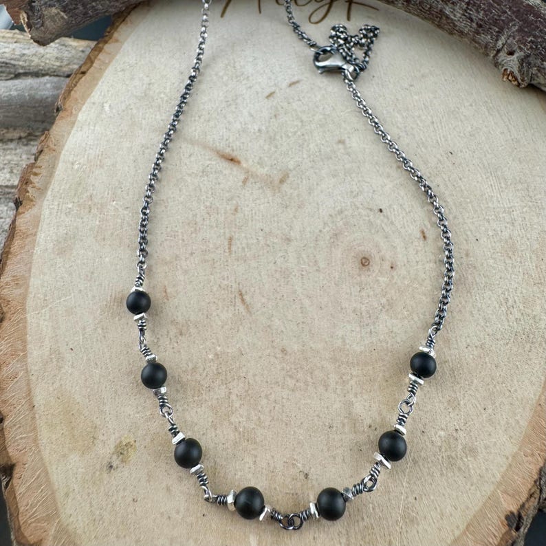 May include: A silver necklace with black beads. The necklace features a delicate chain design with evenly spaced, round black beads. The beads are interspersed with small silver accents, creating a contrast against the dark beads. The necklace is displayed on a wooden surface.