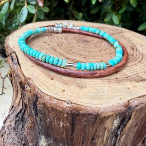 May include: A bracelet featuring a strand of small, round, turquoise-colored beads interspersed with metallic silver-toned spacers, paired with a smooth, dark brown leather cord, secured with a silver-toned lobster claw clasp and metal end caps.