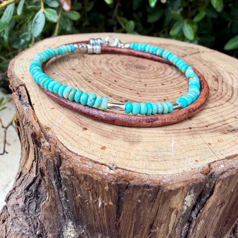 May include: A bracelet featuring a strand of small, round, turquoise-colored beads interspersed with metallic silver-toned spacers, paired with a smooth, dark brown leather cord, secured with a silver-toned lobster claw clasp and metal end caps.