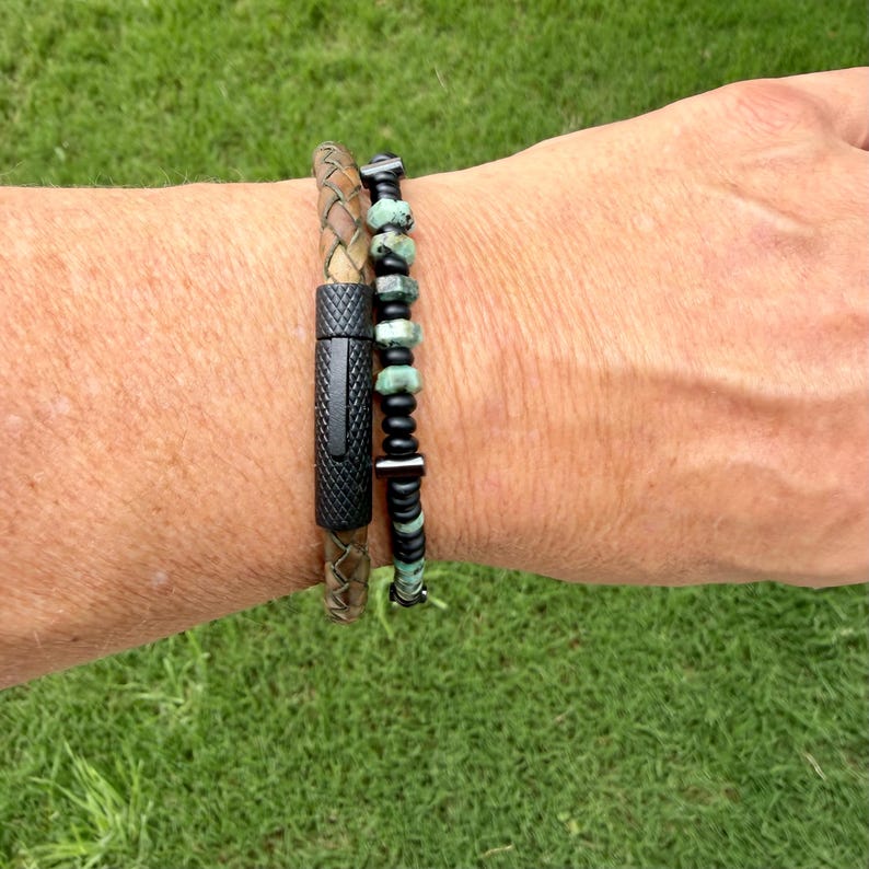 May include: Two bracelets on a wrist. One is a brown leather braided bracelet with a black magnetic clasp. The other is a beaded bracelet with green and black beads. The bracelets are on a person's wrist.