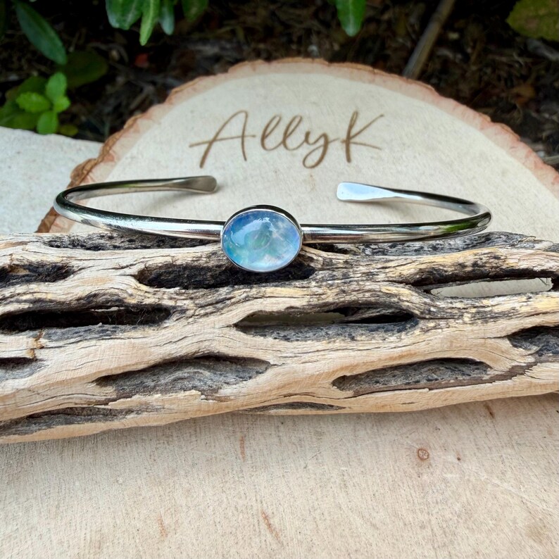 K&ouml;nnte beinhalten: Ein silberfarbenes Armreif-Armband mit einem ovalen, blau-wei&szlig;en Stein im Zentrum. Das Armband wird auf einem Holzst&uuml;ck pr&auml;sentiert, mit dem Namen "Allyk" auf einer Holzscheibe im Hintergrund.