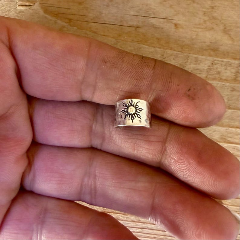 May include: A silver-colored metal ring featuring a hammered texture and a stamped sun design with radiating rays. The band has a wide, open-ended construction with a polished finish, showcasing a detailed celestial motif centered on the front surface.