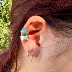 May include: Two silver ear cuffs featuring round, bezel-set turquoise stones with textured metal bands, positioned on the upper ear cartilage, accompanied by two plain, thin silver hoop earrings located in the lower earlobe.