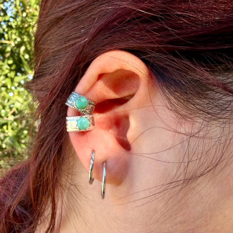 May include: Two silver ear cuffs featuring round, bezel-set turquoise stones with textured metal bands, positioned on the upper ear cartilage, accompanied by two plain, thin silver hoop earrings located in the lower earlobe.