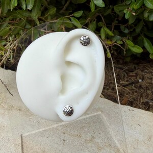 May include: Two circular silver stud earrings featuring a raised cross design. The earrings are displayed on a white ear-shaped model stand. Each stud has a polished metallic finish with a detailed embossed cross emblem centered on the face.