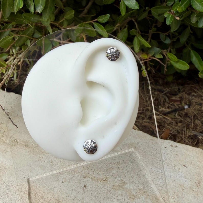 May include: Two circular silver stud earrings featuring a raised cross design. The earrings are displayed on a white ear-shaped model stand. Each stud has a polished metallic finish with a detailed embossed cross emblem centered on the face.