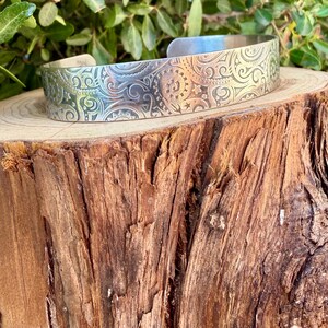 May include: A silver-toned metal cuff bracelet featuring an intricate, embossed pattern of swirling scrolls, small stars, and sun-like motifs. The bracelet has a wide, open-ended band design with a polished, reflective surface finish.