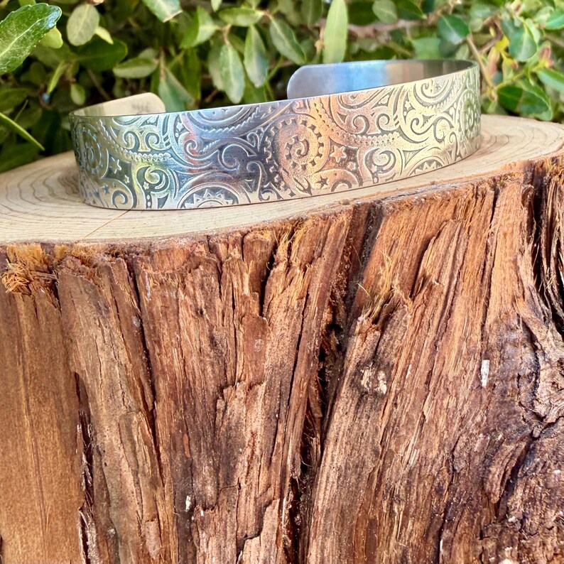 May include: A silver-toned metal cuff bracelet featuring an intricate, embossed pattern of swirling scrolls, small stars, and sun-like motifs. The bracelet has a wide, open-ended band design with a polished, reflective surface finish.