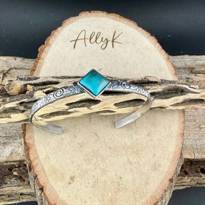 Puede incluir: Una pulsera de plata con una piedra turquesa. La pulsera presenta un dise&ntilde;o floral y el texto "Allyk" sobre un fondo de madera. La piedra turquesa es de forma cuadrada.