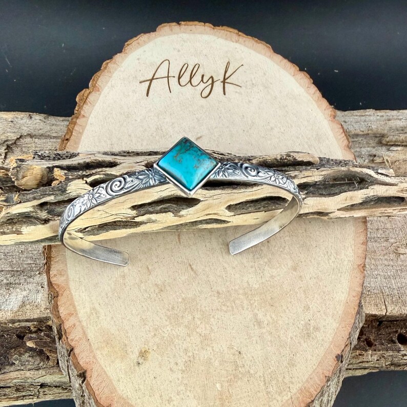 Puede incluir: Una pulsera de plata con una piedra turquesa. La pulsera presenta un dise&ntilde;o floral y el texto "Allyk" sobre un fondo de madera. La piedra turquesa es de forma cuadrada.