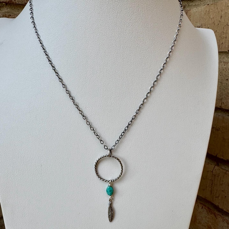 May include: Silver-toned necklace featuring a circular pendant with a turquoise bead and a small feather charm. The chain is a delicate link design. The necklace is displayed on a white mannequin.